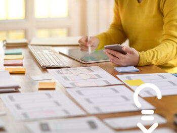 Person sitzt am Schreibtisch und arbeitet am Tablett, während viele Notizen und Zettel um sie herum liegen