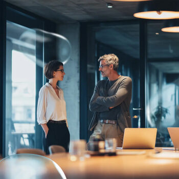 Eine Frau und ein Mann stehen in einem modernen Büro und führen ein Gespräch. Beide scheinen konzentriert zu sein, wobei der Mann seine Arme verschränkt. Laptops und Büromaterialien sind auf einem Tisch im Vordergrund zu sehen.